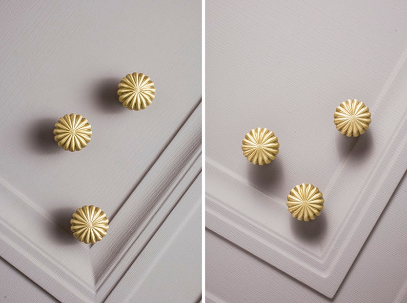 May include: Close-up of several gold-coloured, flower-shaped cabinet knobs. The knobs are arranged on a light grey surface with a textured, paneled design. The knobs have a detailed, ridged pattern.