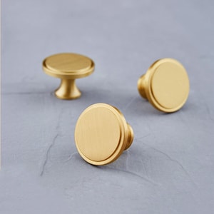 Peut inclure: Trois boutons de meuble dorés avec une finition brossée sont disposés sur une surface grise. Deux boutons ont une face plate et circulaire avec un bord arrondi, tandis que le troisième bouton a une base en piédestal. Les boutons sont probablement pour les meubles.