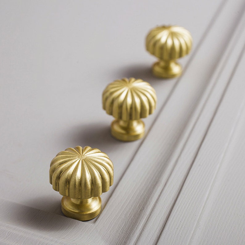 May include: Three gold cabinet knobs with a fluted, mushroom-shaped design are mounted on a light grey cabinet door. The knobs are evenly spaced, creating a symmetrical pattern. The hardware adds a touch of elegance to the furniture.