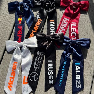 Pode incluir: Uma coleção de laços de cabelo de cetim em várias cores, incluindo branco, preto, vermelho e azul marinho. Cada laço apresenta uma fita longa impressa com logotipos de equipas de corrida e textos como "McLaren", "Red Bull" e "Ferrari".