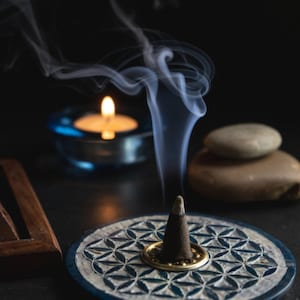 May include: A blue and white ceramic incense holder with a flower of life design. A black incense cone is burning in the holder, with smoke rising from the top.