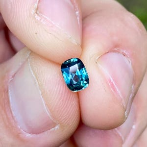 Puede incluir: Una piedra preciosa de corte cojín con una paleta de colores azul verdoso profundo, con una superficie facetada que refleja la luz. La piedra muestra una mezcla de tonos azules oscuros y claros con claridad interna visible y bordes definidos.