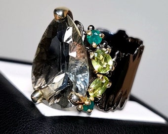 May include: A silver ring with a large pear-shaped clear gemstone and smaller green gemstones. The ring has a textured band.
