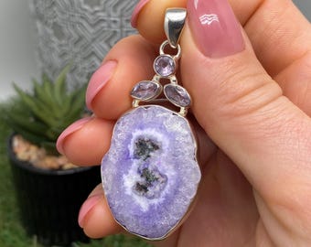 May include: A silver pendant featuring a large, oval-shaped, lavender-colored druzy stone with a dark center. The pendant is accented with three smaller, light purple stones and a silver bail. The pendant is held up by a hand with pink nail polish.