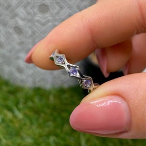 Può includere: Un anello d'argento con un motivo geometrico ripetuto. Ogni forma contiene una piccola gemma rotonda di colore viola chiaro. L'anello è tenuto tra due dita su uno sfondo verde e grigio sfocato.