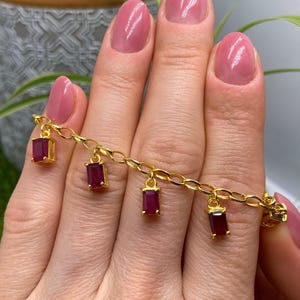 Peut inclure: Un bracelet doré avec un design de chaîne, orné de breloques rectangulaires en rubis. Le bracelet est présenté sur un fond, mettant en valeur les détails et la couleur du bijou. Les breloques en rubis sont rouge foncé.