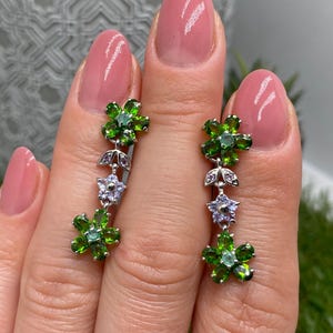 Könnte beinhalten: Silberne Ohrringe mit floralem Design. Jeder Ohrring hat drei grüne Blütengruppen, eine Lavendelblüte und kleine lila Akzente. Die Ohrringe werden von einer Hand mit rosa Nagellack gehalten.