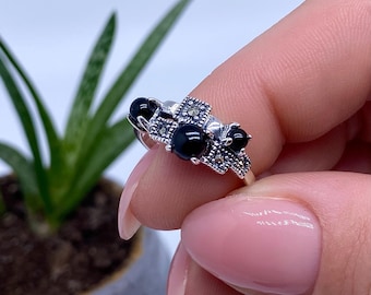 May include: A silver ring featuring three large, round black gemstones and smaller, square marcasite accents. The ring is held up against a white background, showcasing the intricate details of the jewelry.