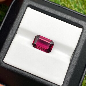 May include: A rectangular, faceted, deep red gemstone, possibly a ruby or garnet, is displayed in a black jewelry box with a white interior. The gem's facets catch the light, showcasing its rich color and clarity. The box is open, revealing the stone.