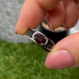 Può includere: Un anello color argento con una pietra preziosa granato rosso scuro dal taglio ovale al centro, circondata da un alone di piccole pietre chiare e arricchita da diverse piccole pietre nere incastonate lungo la fascia su entrambi i lati.