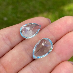 May include: Two pear-shaped, faceted gemstones with a light blue hue. The gems are held in a hand, with a blurred green background. The gems have a clear, transparent appearance, with multiple facets catching the light.