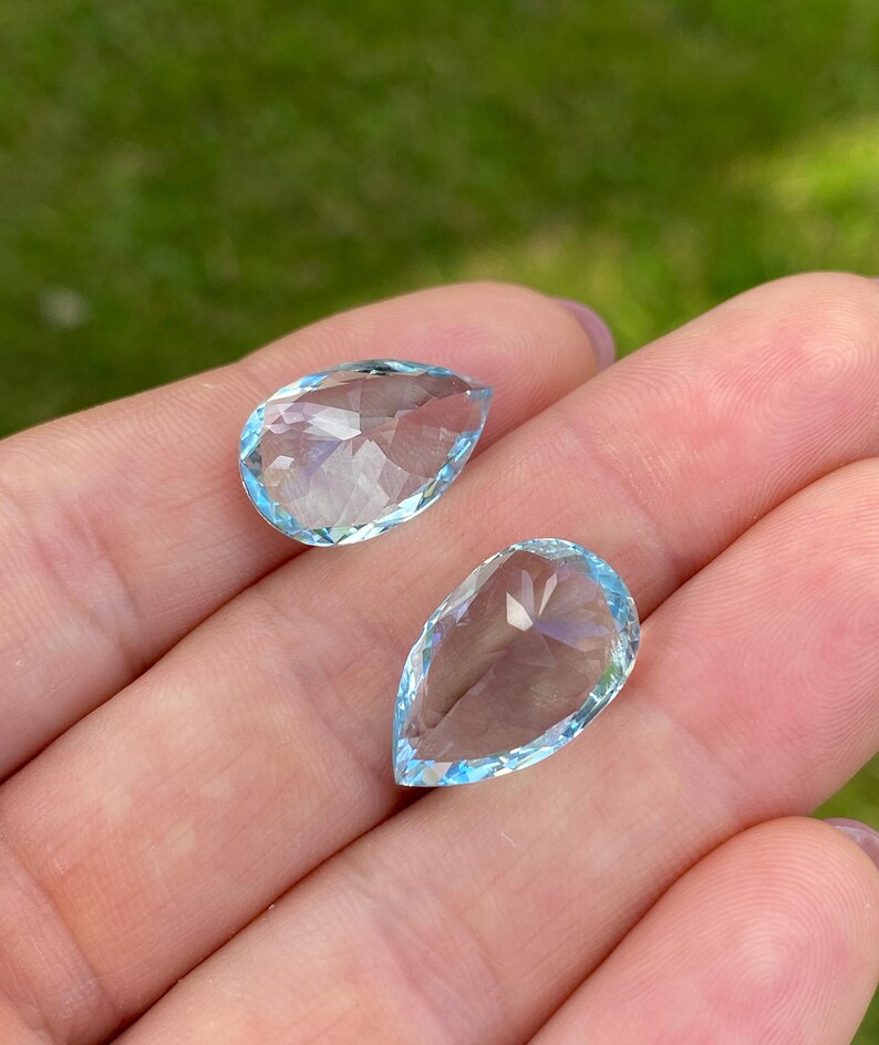 May include: Two pear-shaped, faceted gemstones with a light blue hue. The gems are held in a hand, with a blurred green background. The gems have a clear, transparent appearance, with multiple facets catching the light.