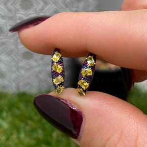 Pode incluir: Um par de brincos curvos com uma fila vertical de quatro pedras preciosas amarelas de corte oval sobre uma base de metal escuro, acentuadas com pequenas pedras pavé de cor vermelho-escuro e azul, criando um contraste vibrante de cores.
