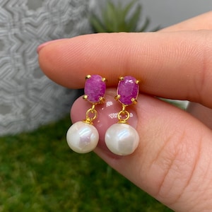 May include: A pair of gold drop earrings featuring oval-shaped, faceted pink ruby gemstones at the top, connected by small gold loops to round, white, lustrous pearls hanging at the bottom of each earring.