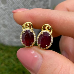 May include: A pair of gold stud earrings featuring oval-cut deep red rubies surrounded by a halo of small white diamonds. Each earring has a decorative gold swirl accent at the top, securing the gemstone in a prong setting.