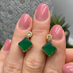 Op de afbeelding: Een paar gouden oorbellen met vierkant geslepen groene edelstenen die aan kleinere ronde knopjes hangen. Elk knopje is versierd met een halo van kleine witte steentjes rondom een centrale lichtgroene steen in een gouden zetting.
