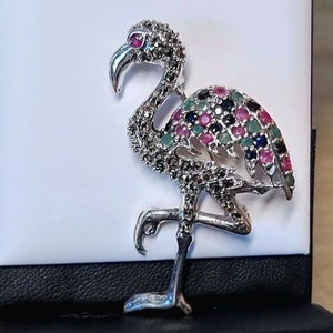 Può includere: Un fermaglio a forma di fenicottero in argento con un design dettagliato. Il fenicottero è adornato con pietre preziose colorate, tra cui rosa, verde e blu.