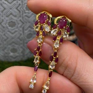 Puede incluir: Un par de pendientes colgantes en tono dorado con rubíes de color rosa intenso en talla ovalada y piedras blancas transparentes en forma de pera, dispuestas en un patrón floral en cascada, con pequeñas piedras redondas en las puntas.