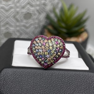 Può includere: Un anello d'argento con un design a forma di cuore. Il cuore è riempito con un mosaico di pietre preziose colorate, tra cui rosa, blu, giallo e verde. La fascia dell'anello è decorata con piccole pietre preziose rosse.