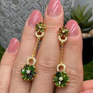 Könnte beinhalten: Ein Paar goldene Ohrhänger mit einem blumenförmigen Stecker, der einen grünen Edelstein und einen kleinen klaren Stein in der Mitte aufweist. Eine feine Goldkette verbindet den Stecker mit einem größeren blumenförmigen Anhänger mit dunkelgrünen Blütenblättern.