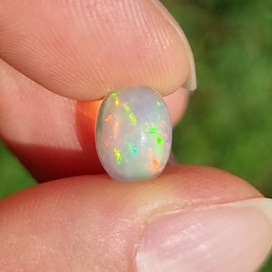 May include: A white opal gemstone with a light gray body color and iridescent flashes of green, orange, and red.