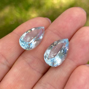 May include: Two pear-shaped, faceted aquamarine gemstones. The gems are a light blue color and are held in a hand against a blurred green background. The facets catch the light, creating a sparkling effect.