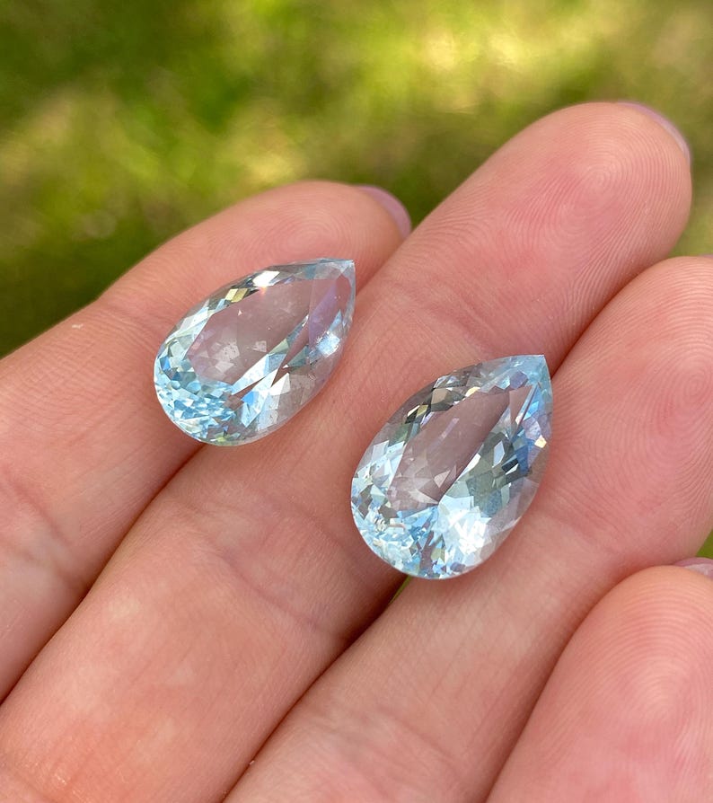 May include: Two pear-shaped, faceted aquamarine gemstones. The gems are a light blue color and are held in a hand against a blurred green background. The facets catch the light, creating a sparkling effect.