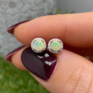 Pode incluir: Um par de brincos redondos com opalas iridescentes centrais rodeadas por um halo de pequenas pedras transparentes cravadas numa armação de metal prateado. As opalas exibem flashes de luz verde, laranja e amarela nas cravações.