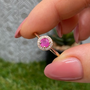 May include: A gold ring featuring a round, bright pink gemstone surrounded by a halo of smaller, clear stones. The ring's band is thin and polished, and the setting is delicate. The ring is held up against a soft background.