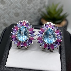 May include: A pair of silver-toned earrings featuring a central pear-shaped light blue gemstone surrounded by a halo of marquise-cut purple, light blue, and pink gemstones arranged in a floral pattern, presented in a black display box.