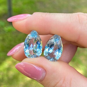 May include: Two pear-shaped, faceted aquamarine gemstones. The gems are a light, translucent blue with internal reflections. The stones are held between fingers with pink nail polish.