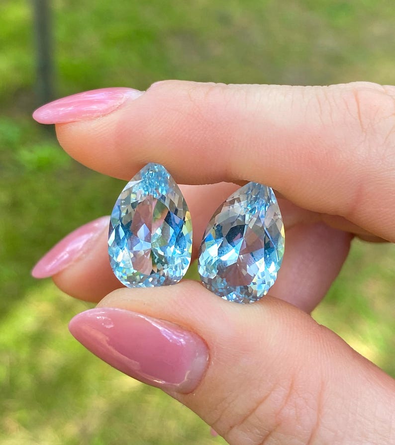 May include: Two pear-shaped, faceted aquamarine gemstones. The gems are a light, translucent blue with internal reflections. The stones are held between fingers with pink nail polish.