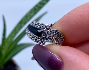 May include: A silver ring with a black onyx stone and marcasite accents. The ring has a geometric design with a triangular onyx center stone and a band adorned with small, sparkling marcasite stones. The ring is held up by a person's fingers.