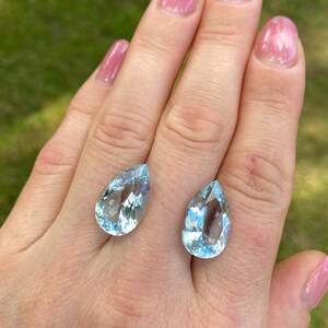 May include: Two pear-shaped, light blue gemstones with internal facets. The gems are displayed on a hand with pink nail polish, set against a blurred green background. The stones are likely aquamarine.