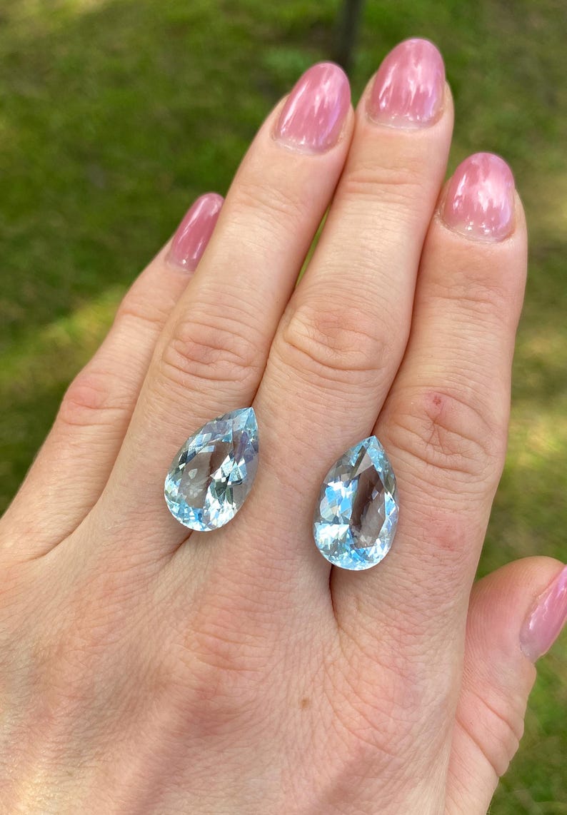 May include: Two pear-shaped, light blue gemstones with internal facets. The gems are displayed on a hand with pink nail polish, set against a blurred green background. The stones are likely aquamarine.