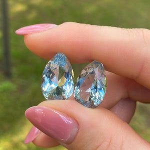 May include: Two pear-shaped, faceted gemstones with a light blue hue. The gems are held between fingers with a blurred green background. The gems have multiple facets that reflect light, creating a sparkling effect. The gems are likely aquamarine.