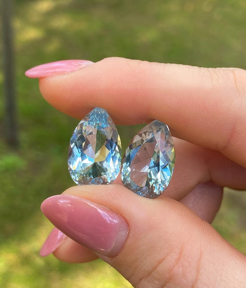 May include: Two pear-shaped, faceted gemstones with a light blue hue. The gems are held between fingers with a blurred green background. The gems have multiple facets that reflect light, creating a sparkling effect. The gems are likely aquamarine.