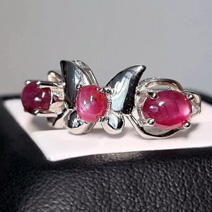 Könnte beinhalten: Ein silberner Ring mit einem Schmetterlingsdesign, besetzt mit drei ovalen, leuchtend roten Edelsteinen. Band und Schmetterlingsdetails des Rings sind aus poliertem Silber gefertigt, was einen auffälligen Kontrast zu den rubinroten Steinen bildet.