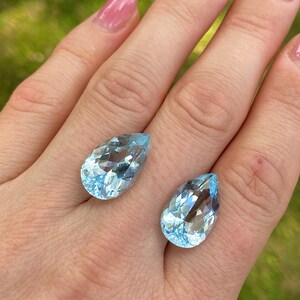 May include: Two pear-shaped, light blue gemstones with facets, displayed on a hand. The gems have a clear, crystalline appearance, reflecting light. The background is a blurred green, suggesting an outdoor setting. The gems are likely aquamarine.