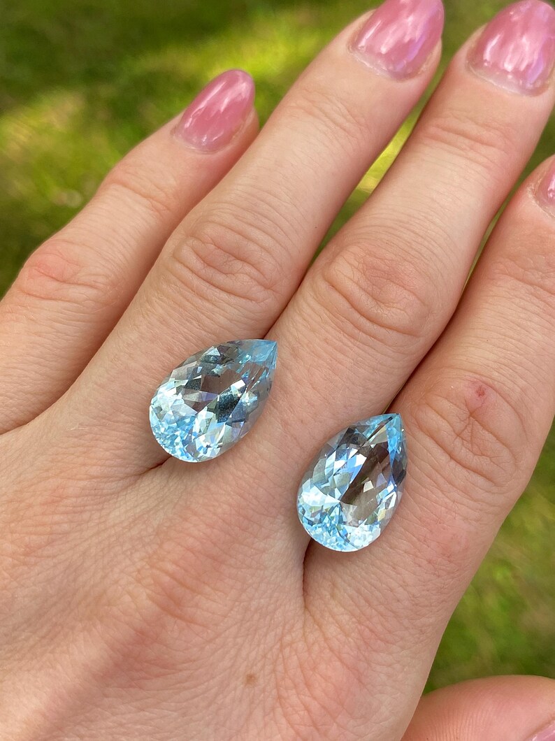 May include: Two pear-shaped, light blue gemstones with facets, displayed on a hand. The gems have a clear, crystalline appearance, reflecting light. The background is a blurred green, suggesting an outdoor setting. The gems are likely aquamarine.