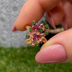 Puede incluir: Anillo de oro rosa con diseño floral y piedras preciosas rojas, verdes y amarillas. Los intrincados detalles del anillo incluyen acentos en forma de hojas alrededor de las piedras de colores.