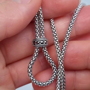 Könnte beinhalten: Nahaufnahme einer silbernen Halskette mit einer strukturierten, perlenbesetzten Kette. Die Halskette hat eine dekorative, verschiebbare Perle mit dunklen Akzenten. Die Kette ist geschlungen und zeigt das Design und die Textur des Schmucks.