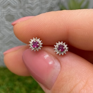May include: A pair of stud earrings featuring a central round-cut deep pink ruby surrounded by a halo of small, clear, sparkling white stones set in a polished silver-toned metal frame, creating a floral-inspired circular design.