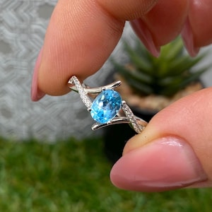 Pode incluir: Um anel de prata com uma grande pedra preciosa oval azul brilhante. A banda do anel tem um design delicado e torcido com pequenas pedras claras de destaque. O anel é segurado contra um fundo verde desfocado.