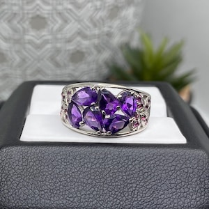 Op de afbeelding: Een zilverkleurige ring met een cluster van peervormige paarse amethist edelstenen in het midden, geaccentueerd door kleinere ronde roze stenen aan de zijkanten. De ringband heeft een open, decoratief metalen ontwerp rond de stenen.