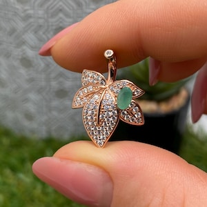 Puede incluir: Colgante con forma de hoja en oro rosa, adornado con pequeñas piedras preciosas transparentes y una gema verde ovalada en el centro. El diseño de la hoja presenta detalles intrincados.