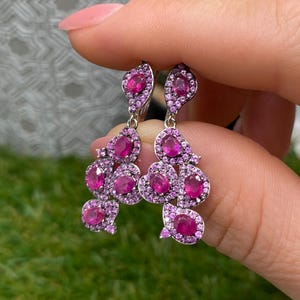 Könnte beinhalten: Ein Paar Ohrhänger mit mehreren ovalen rosa Steinen, die von kleinen, runden rosa Pavé-Kristallen umgeben sind und in einer silberfarbenen Metallfassung sitzen. Das Design besteht aus einem oberen Stein und einem größeren, traubenförmigen Unterteil.