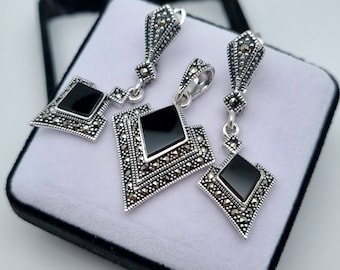 May include: A silver-toned jewelry set featuring a pendant and matching earrings. Each piece has a black, diamond-shaped center stone surrounded by small, sparkling accents. The set is displayed in a black box with a white interior.