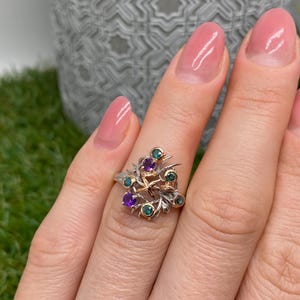 Pode incluir: Anel de prata e ouro com design floral e pedras preciosas roxas e verdes. O anel apresenta um motivo detalhado inspirado na natureza com um grupo de pedras. As pedras são engastadas em biséis de ouro.