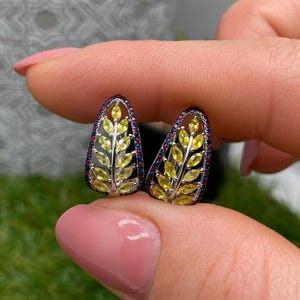 May include: A pair of silver leaf-shaped earrings. Each earring has a central design of yellow gemstones in a leaf pattern, framed by blue and red gemstones. The earrings are held up by a person's fingers.